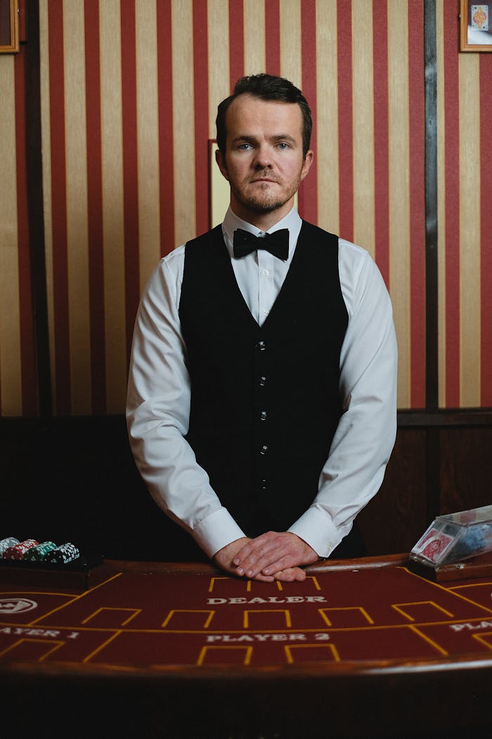 A professional casino croupier in formal attire stands ready at the roulette table.
